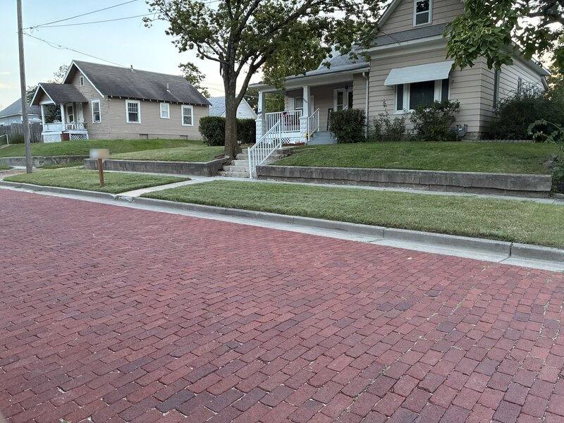 A mowed green lawn and a stone retaining wall running along a brick-paved Wichita street, with two older houses and a large tree behind it.